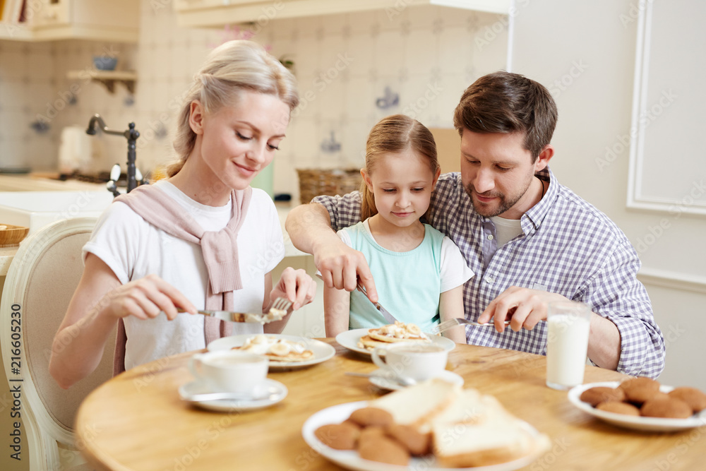 Happy content friendly family in casual clothing eating breakfast in morning: young father assisting daughter to cut pancakes in dining room Happy content friendly family in casual clothing eating breakfast in morning: young father assisting daughter to cut pancakes in dining room