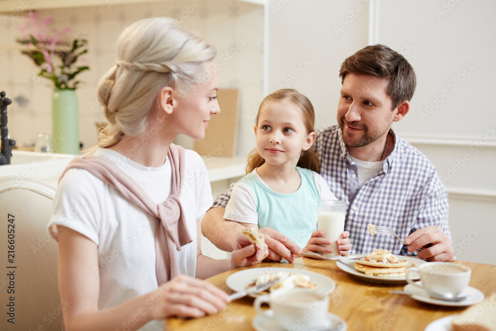 Content beautiful family sitting at dining table and chatting while eating delicious homemade pancakes in domestic kitchen Content beautiful family sitting at dining table and chatting while eating delicious homemade pancakes in domestic kitchen