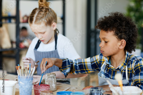 Serious creative schoolboy putting paintbrush into jar with paint while sitting at lesson of arts and crafts