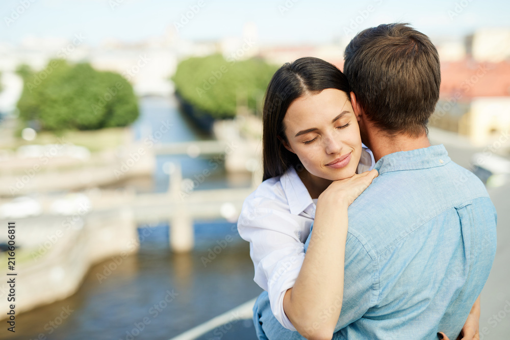 Calm attractive young lady with straight black hair keeping eyes closed and hugging with tenderness boyfriend while they standing on roof in city