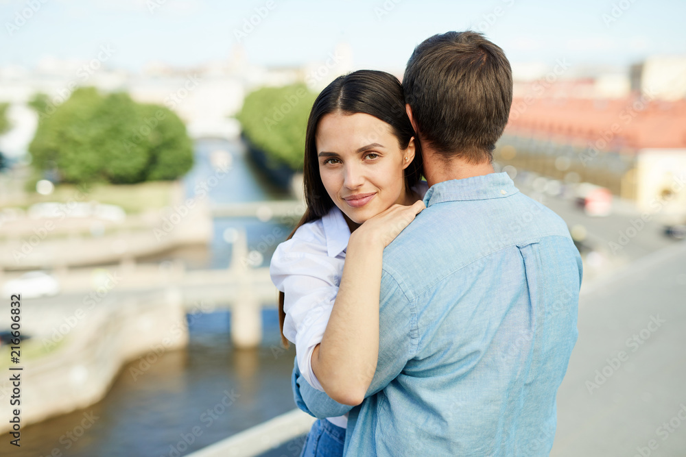 Content attractive brunette girl embracing boyfriend and looking at camera while they standing on city roof during romantic date