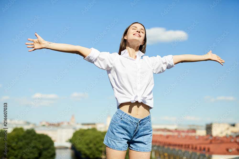 Happy free young woman in white shirt and denim shorts outstretching arms and keeping eyes closed while standing on roof in city, freedom concept