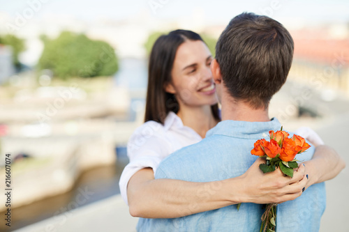 Focus on small bouquet of orange roses in hands of girlfriend, happy affectionate woman falling in love with boyfriend cuddling him on roof