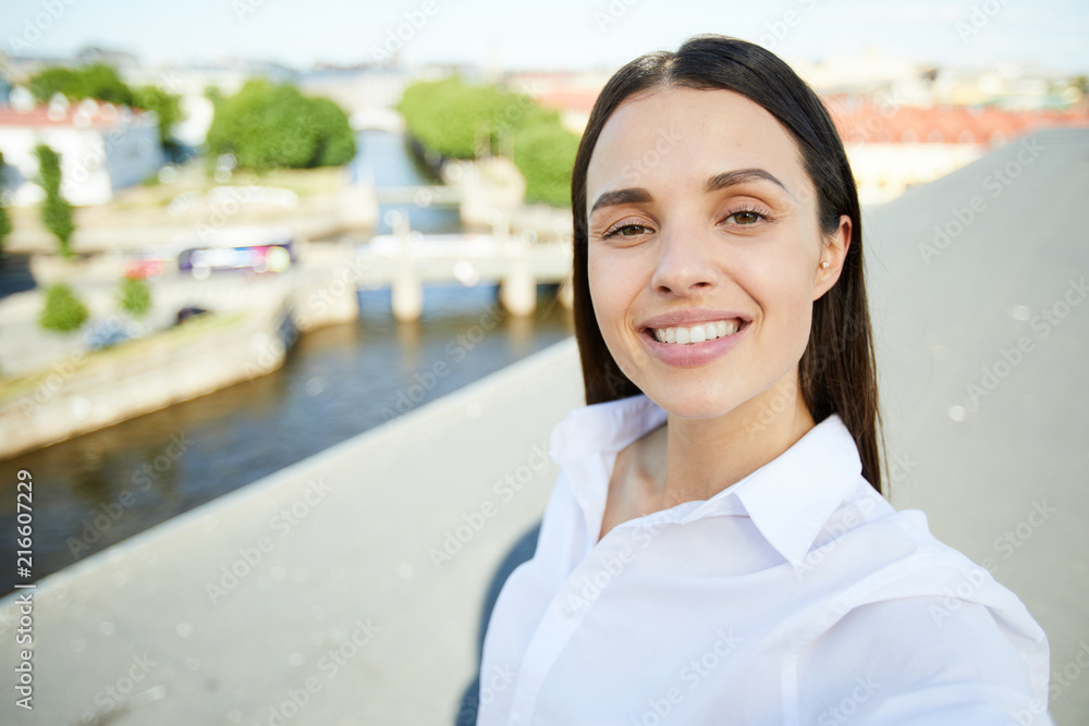 Cheerful pretty young brunette lady with beautiful smile taking selfie against city while standing on roof