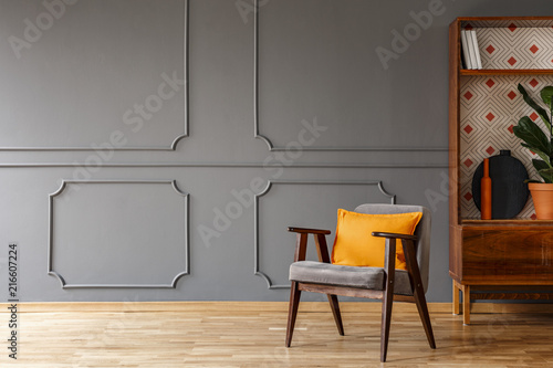 Grey armchair with orange pillow standing in real photo of dark grey living room interior with wainscoting on the wall and empty space for your coffee table photo