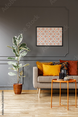 Real photo of poster with geometric pattern hanging on the wall with wainscoting in dark living room interior with fresh plant, sofa with cushions and metal end table with decor