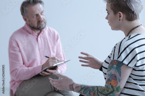A troubled teenage girl with tattoos showing her emotions and explaining her situation and actions to a psychotherapist during an individual therapy meeting.