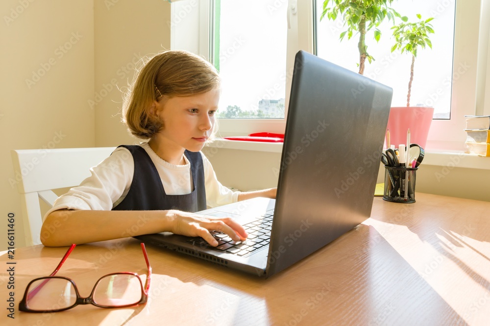 Little schoolgirl uses computer sitting at a desk at home. School, education, knowledge and children. Little schoolgirl uses computer sitting at a desk at home. School, education, knowledge and children.