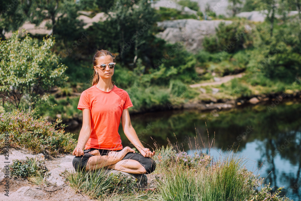 Young girl in pink T-shirt and black leggings sitting on a stone in lotus  pose near the lake. concept of yoga, pilates, healthy life. Stock Photo |  Adobe Stock