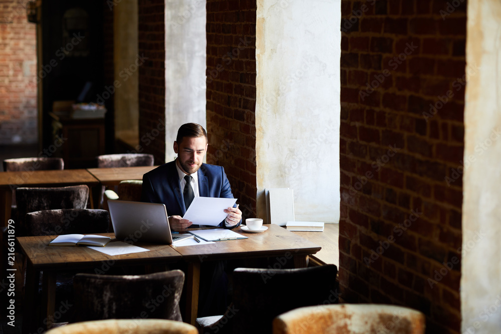 Content young bearded businessman satisfied with results viewing report data while working in restaurant with loft interior Content young bearded businessman satisfied with results viewing report data while working in restaurant with loft interior