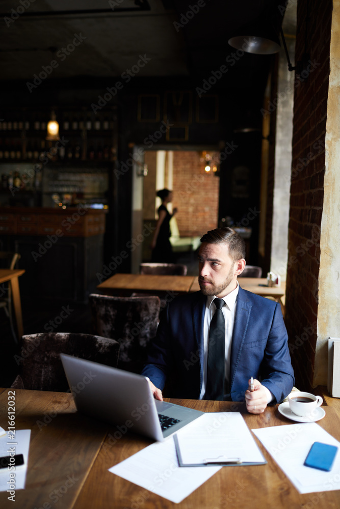 Serious confident brutal businessman using laptop while working in modern restaurant, he looking away while connecting laptop to wifi Serious confident brutal businessman using laptop while working in modern restaurant, he looking away while connecting laptop to wifi