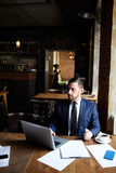 Serious confident brutal businessman using laptop while working in modern restaurant, he looking away while connecting laptop to wifi