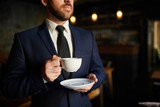 Close-up of serious pensive businessman in formal suit standing and holding dish while drinking coffee during break
