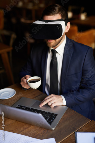 Serious young businessman in VR headset using laptop and interactive simulator for work while drinking coffee in restaurant
