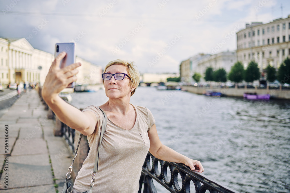 Aged lady in glasses leaning on fence near river and posing for selfie on street of beautiful city