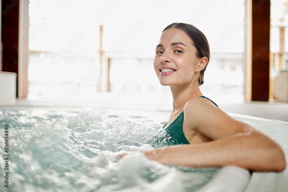 Young woman relaxing in soft waving water while sitting in jacuzzi bath at spa center - obrazy, fototapety, plakaty 