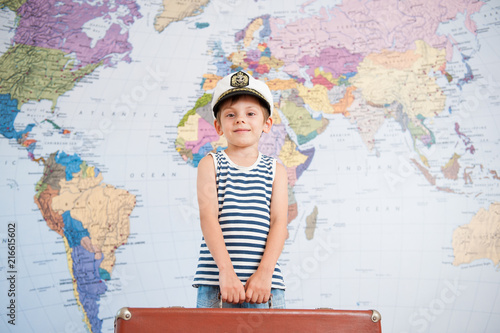 happy smiling kid in captain cap holding heavy suitcase on world map background