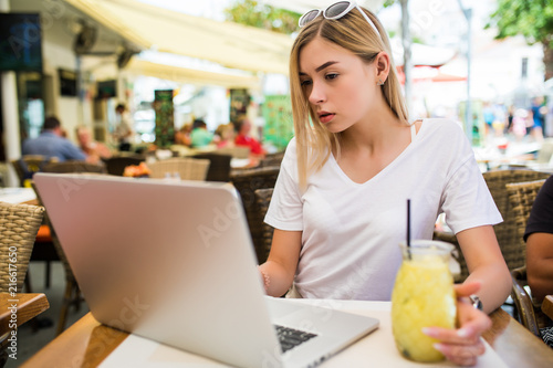 Young woman sits in front of opened laptop computer, drinks fresh summer cocktail, enjoys online communication and free internet