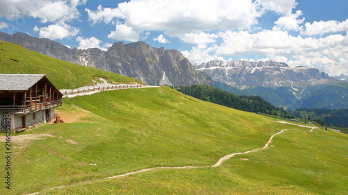 Puez Odle mountain range (on the left) and Sella Group mountains (on the right) viewed from a hiking path near Raiser Pass with a mountain hut in the foreground, Val Gardena, Dolomites, Italy