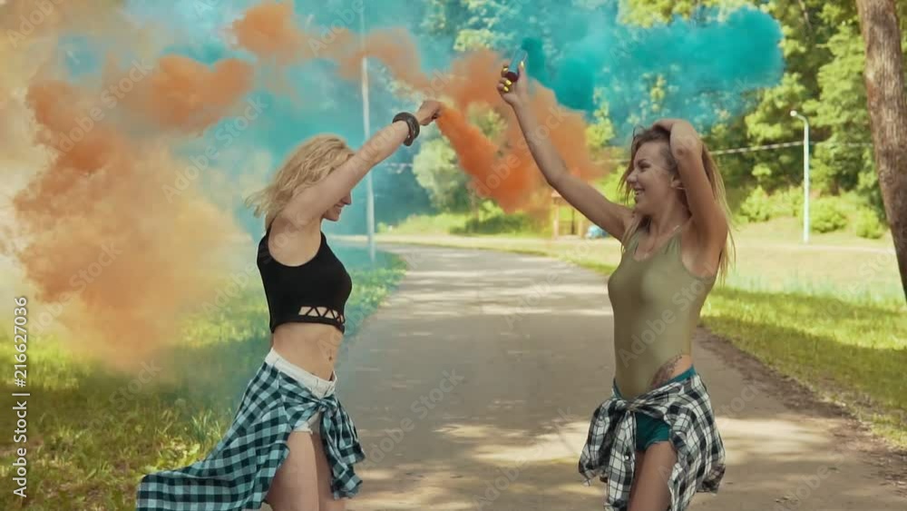 Beautiful hipster girls in beautiful clothes runs in park waving colored smoke. Young girls have fun and jump outdoors in slow motion. Party concept