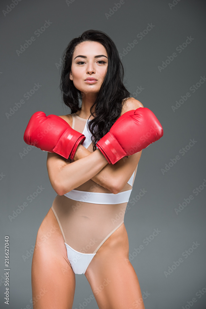attractive sportive woman in white swimwear standing with crossed arms in boxing gloves isolated on grey
