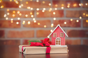 Christmas gift box and wooden toy house on background with fairy lights in bokeh. Holiday season