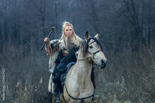 Blonde warrior viking woman riding horse with ax in hand against forest background ready to attack.