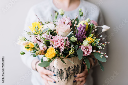 Fototapeta Naklejka Na Ścianę i Meble -  Very nice florist woman holding a big beautiful fresh flower bouquet of narcissus, hyacinth, eustoma,roses in yellow and pastel pink colors on the grey wall background, close up view