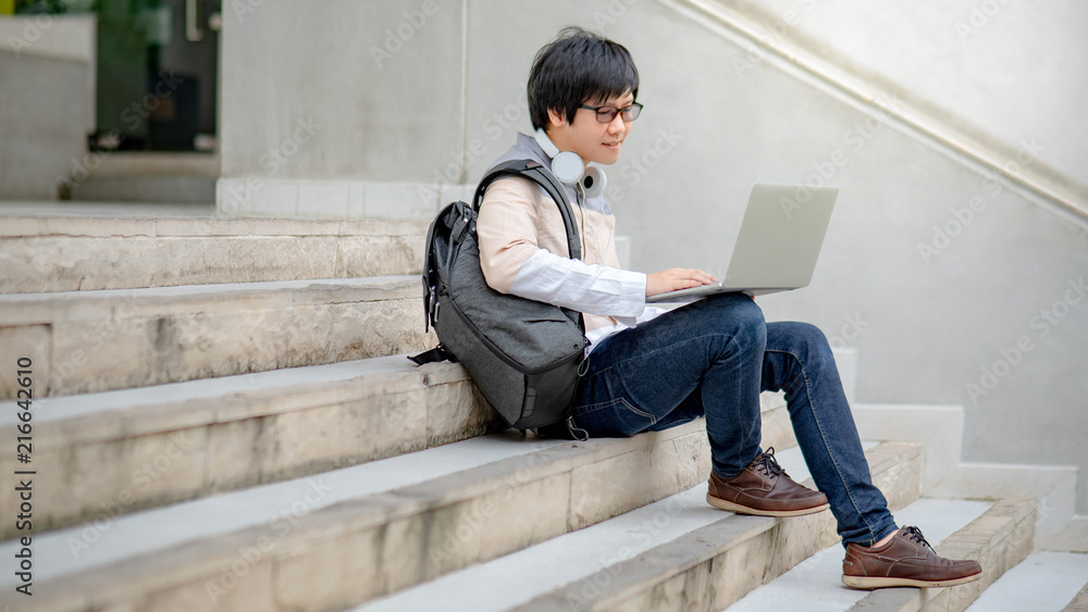 Young Asian man sitting on stair working with laptop computer in college building. Male university student or high school lifestyle. Education concept