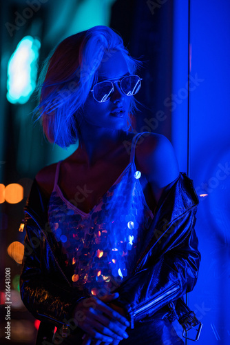 beautiful young woman in glossy tank top and sunglasses on street at night under blue light