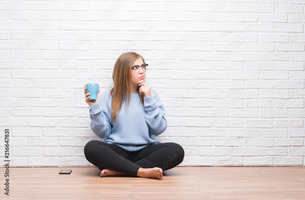 Young adult woman sitting on the floor over white brick wall driking cup of coffee serious face thinking about question, very confused idea Young adult woman sitting on the floor over white brick wall driking cup of coffee serious face thinking about question, very confused idea