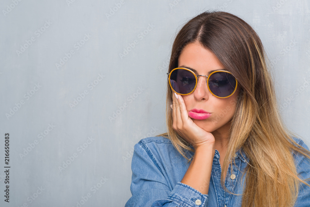 Young adult woman over grunge grey wall wearing retro sunglasses thinking looking tired and bored with depression problems with crossed arms. Young adult woman over grunge grey wall wearing retro sunglasses thinking looking tired and bored with depression problems with crossed arms.