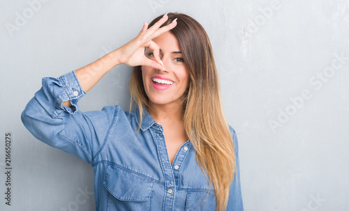 Young adult woman over grey grunge wall wearing denim outfit with happy face smiling doing ok sign with hand on eye looking through fingers
