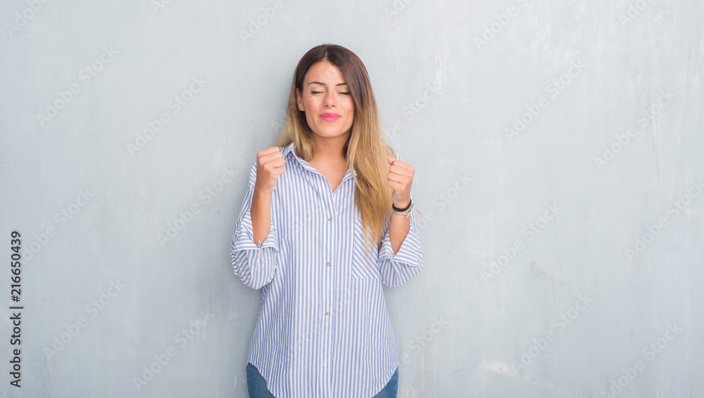 Young adult woman over grey grunge wall wearing fashion business outfit excited for success with arms raised celebrating victory smiling. Winner concept. Young adult woman over grey grunge wall wearing fashion business outfit excited for success with arms raised celebrating victory smiling. Winner concept.