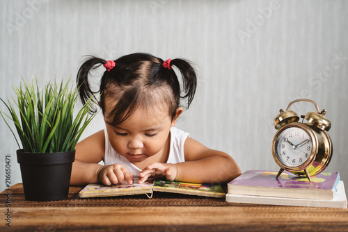 asian baby toddler reading book on wooden table with alarm clock. concept of early education, child learning and development