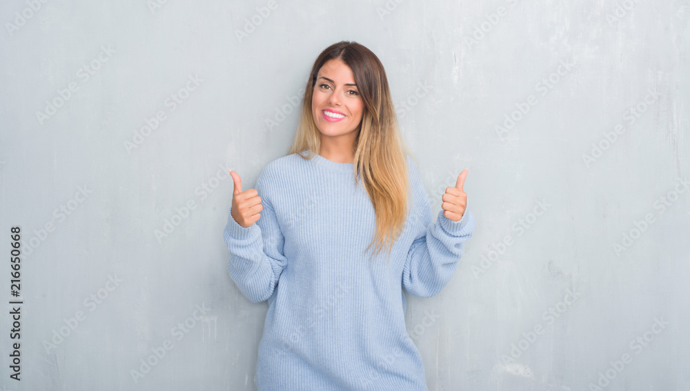 Young adult woman over grey grunge wall wearing winter outfit success sign doing positive gesture with hand, thumbs up smiling and happy. Looking at the camera with cheerful expression, winner Young adult woman over grey grunge wall wearing winter outfit success sign doing positive gesture with hand, thumbs up smiling and happy. Looking at the camera with cheerful expression, winner