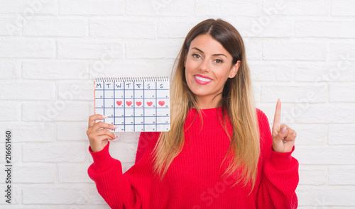 Young adult woman over white brick wall holding period calendar surprised with an idea or question pointing finger with happy face, number one
