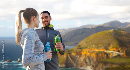 fitness, sport and people concept - smiling couple with bottles of water over bixby creek bridge on big sur coast of california background