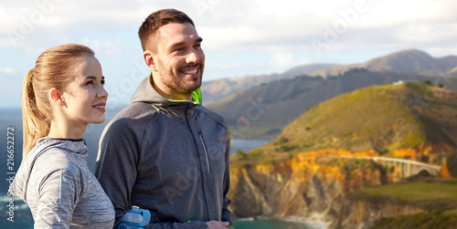 fitness, sport and people concept - smiling couple with bottles of water over bixby creek bridge on big sur coast of california background