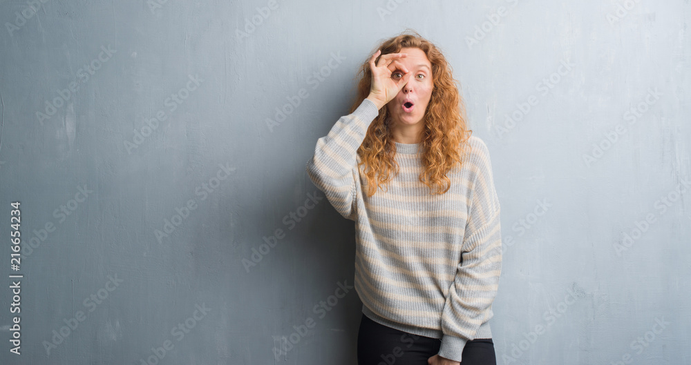 Young redhead woman over grey grunge wall doing ok gesture shocked with surprised face, eye looking through fingers. Unbelieving expression. Young redhead woman over grey grunge wall doing ok gesture shocked with surprised face, eye looking through fingers. Unbelieving expression.