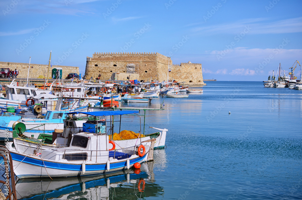 Heraklion, Crete Island - Greece. Traditional colorful fishing boats are docked at the old port of Heraklion city in front of the fortress Koules (castello a mare). Sunny day, blue cloudy sky