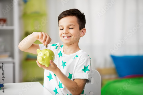 money, finances, childhood and people concept - smiling little boy putting coin into piggy bank at home