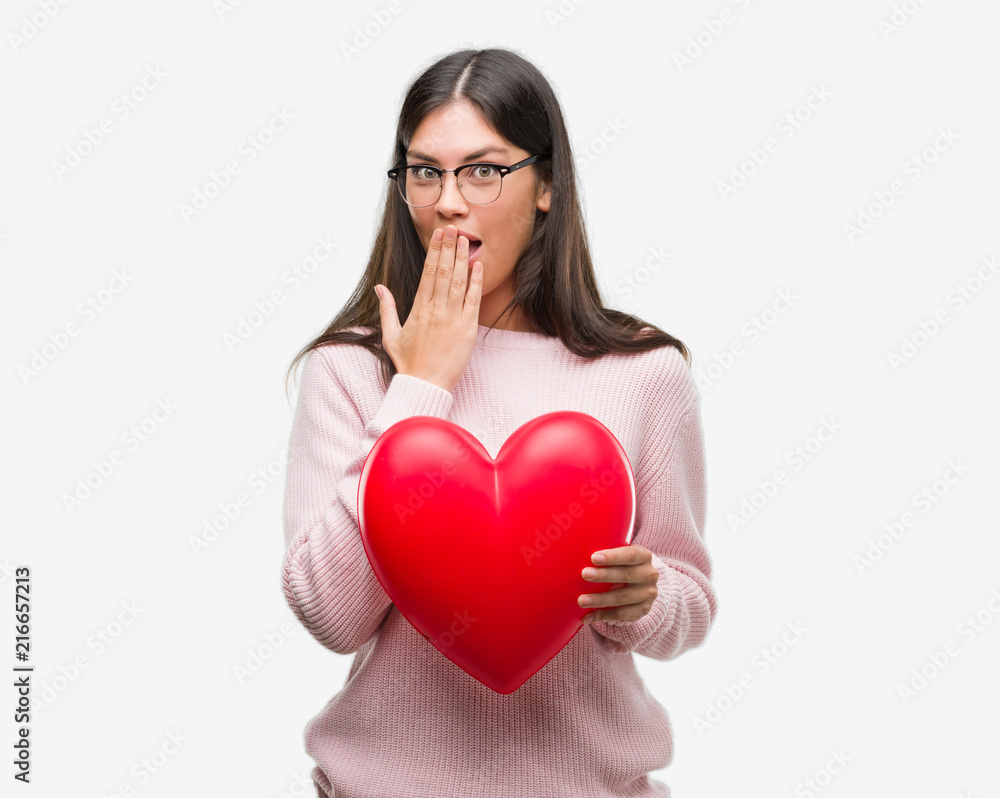 Young hispanic woman holding heart in love cover mouth with hand shocked with shame for mistake, expression of fear, scared in silence, secret concept
