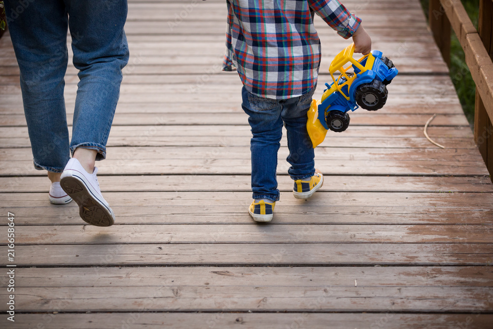 Child with a toy car. Back view on toddler boy in plaid shirt and blue jeans running on wooden surface in the park. Child with a toy car. Back view on toddler boy in plaid shirt and blue jeans running on wooden surface in the park.