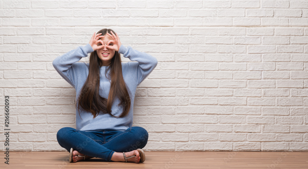 Young Chinese woman sitting on the floor over brick wall doing ok gesture like binoculars sticking tongue out, eyes looking through fingers. Crazy expression. Young Chinese woman sitting on the floor over brick wall doing ok gesture like binoculars sticking tongue out, eyes looking through fingers. Crazy expression.
