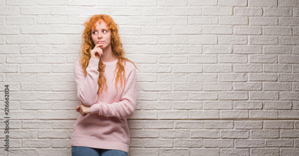 Young redhead woman standing over brick wall with hand on chin thinking about question, pensive expression. Smiling with thoughtful face. Doubt concept. Young redhead woman standing over brick wall with hand on chin thinking about question, pensive expression. Smiling with thoughtful face. Doubt concept.