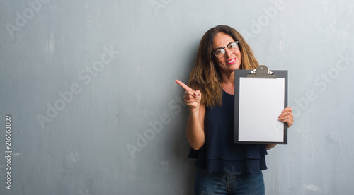 Middle age hispanic woman standing over grey grunge wall holding clipboard very happy pointing with hand and finger to the side