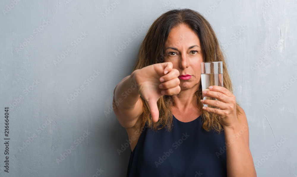 Middle age hispanic woman drinking glass of water with angry face, negative sign showing dislike with thumbs down, rejection concept Middle age hispanic woman drinking glass of water with angry face, negative sign showing dislike with thumbs down, rejection concept