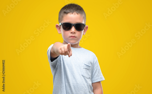 Dark haired little child wearing sunglasses pointing with finger to the camera and to you  hand sign  positive and confident gesture from the front