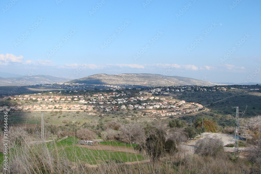 The landscape of small towns in the central part of Israel at the foot of low hills in the background of a cloudy blue sky.